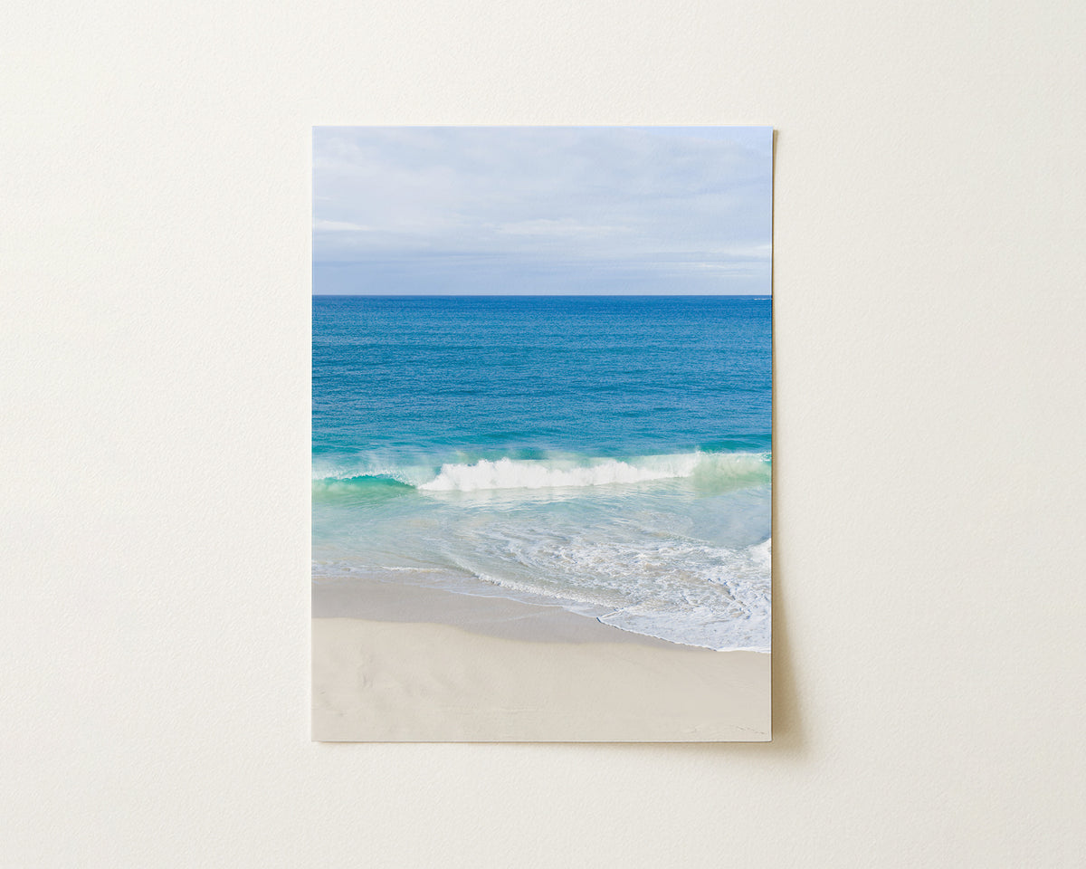 Fine art paper print of aerial photography of Alkimos Beach, a small wave rolling onto pale sand beneath a blue sea and cloudy sky in portrait orientation