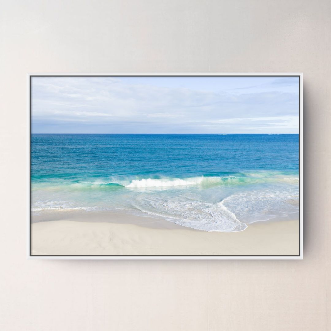White framed canvas print of aerial photography of Alkimos Beach, a small wave rolling onto pale sand beneath a blue sea and cloudy sky