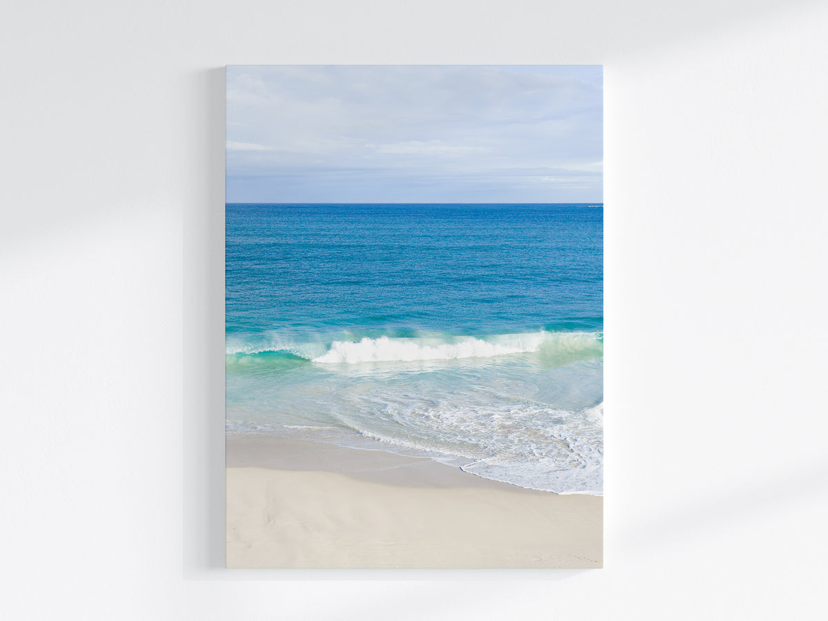 Stretched canvas print of aerial photography of Alkimos Beach, a small wave rolling onto pale sand beneath a blue sea and cloudy sky in portrait orientation on a wall