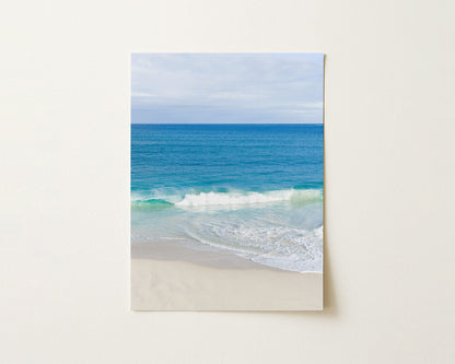 Fine art paper print of aerial photography of Alkimos Beach, a small wave rolling onto pale sand beneath a blue sea and cloudy sky in portrait orientation