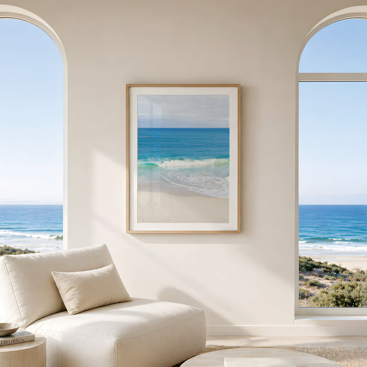 Framed fine art print of a wave rolling onto pale sand beneath deep blue ocean, hanging in a living room with arched windows and a sea view