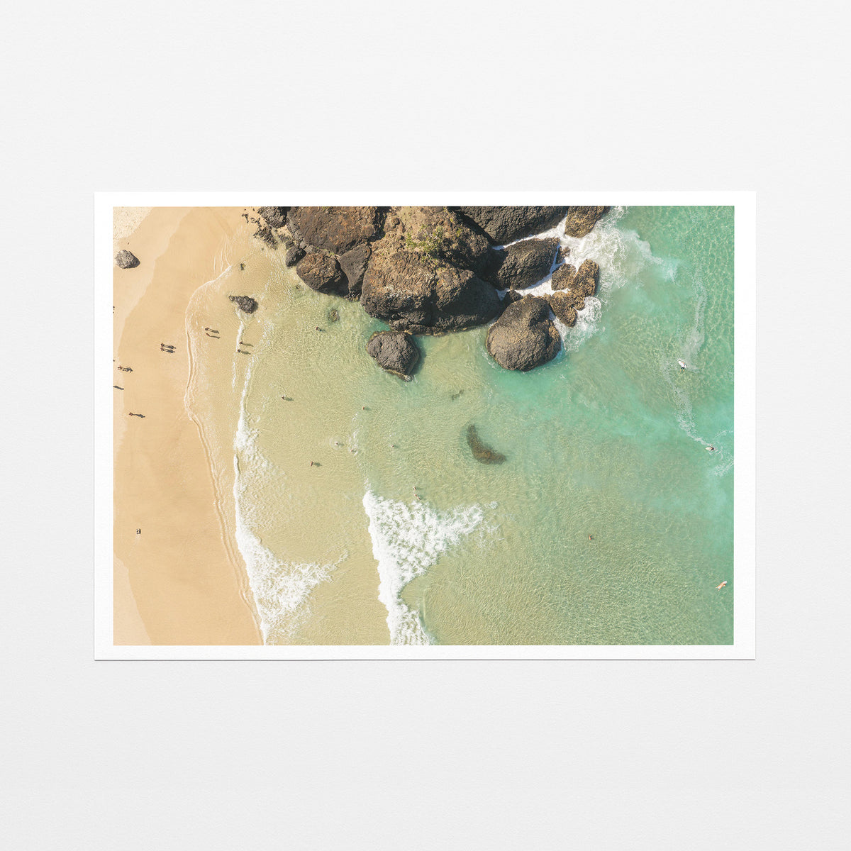 Fine art paper print of aerial photography of Dreamtime Beach, a dark rock outcrop beside golden sand and green water with a white border