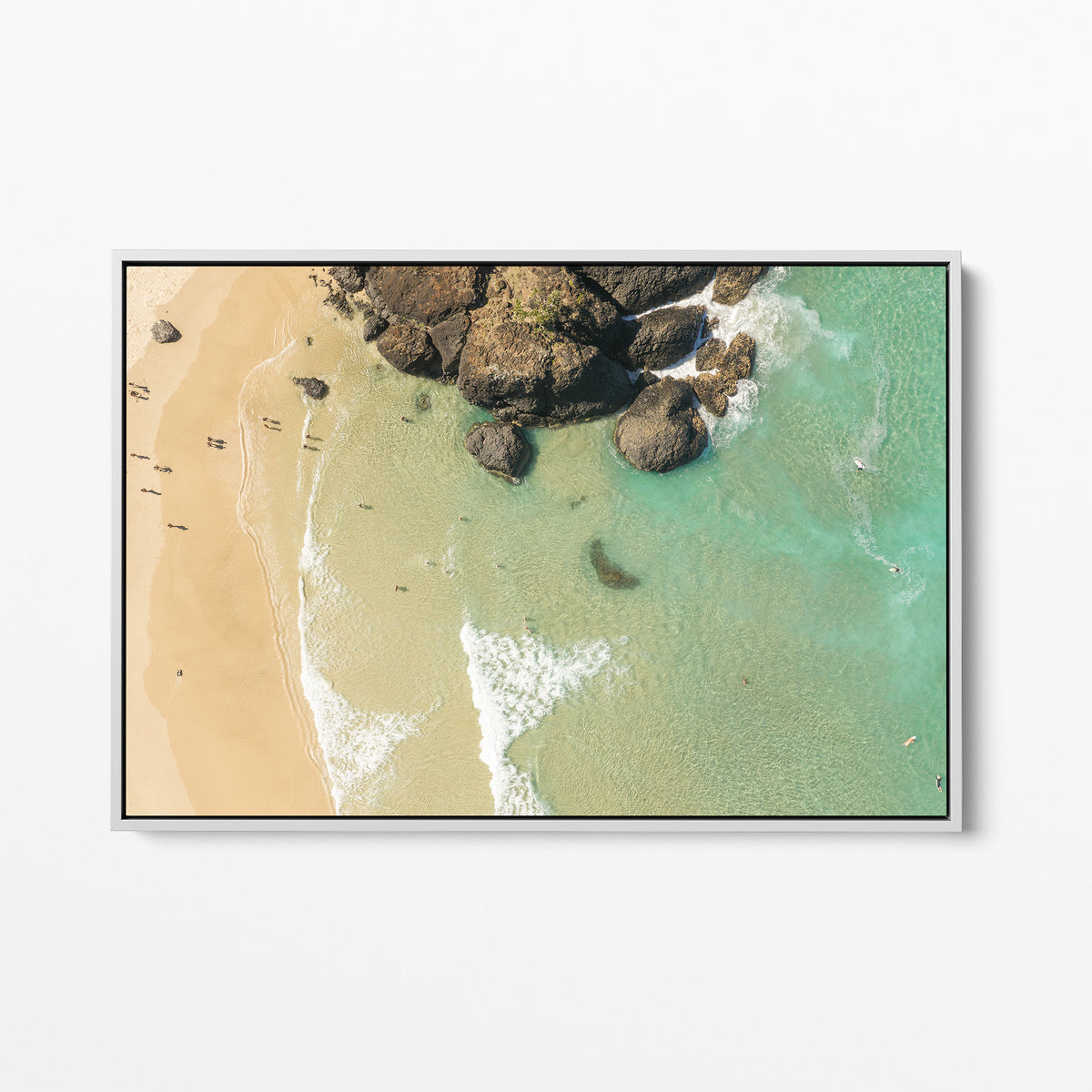 White framed canvas print of aerial photography of Dreamtime Beach, a dark rock outcrop beside golden sand and green water