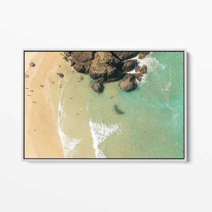 White framed canvas print of aerial photography of Dreamtime Beach, a dark rock outcrop beside golden sand and green water