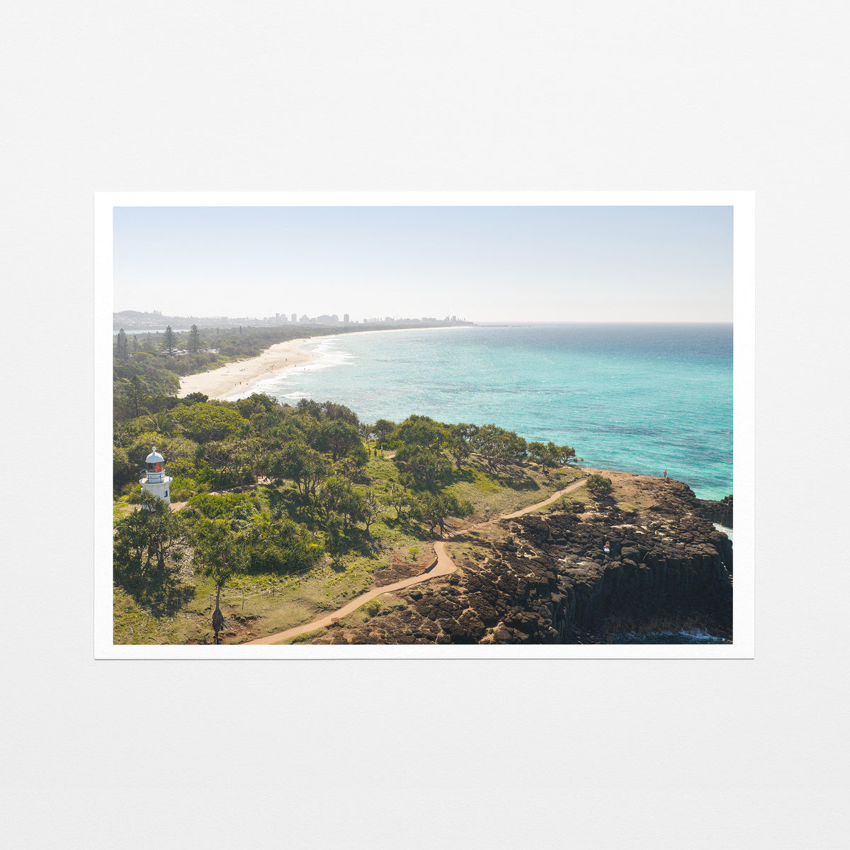 Fine art paper print of aerial photography of Fingal Head Lighthouse, bushland with a dirt track, a rocky cliff, the beach and turquoise sea with a white border